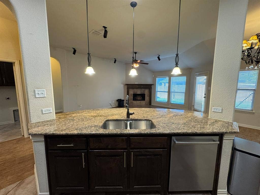 3953 Hollow Lake Road, Fort Worth, TX 76262 Kitchen island featuring stone-surfaced countertops, stainless steel undermount double-basin sink, dark wood cabinetry, three pendant light fixtures, and a stainless steel dishwasher