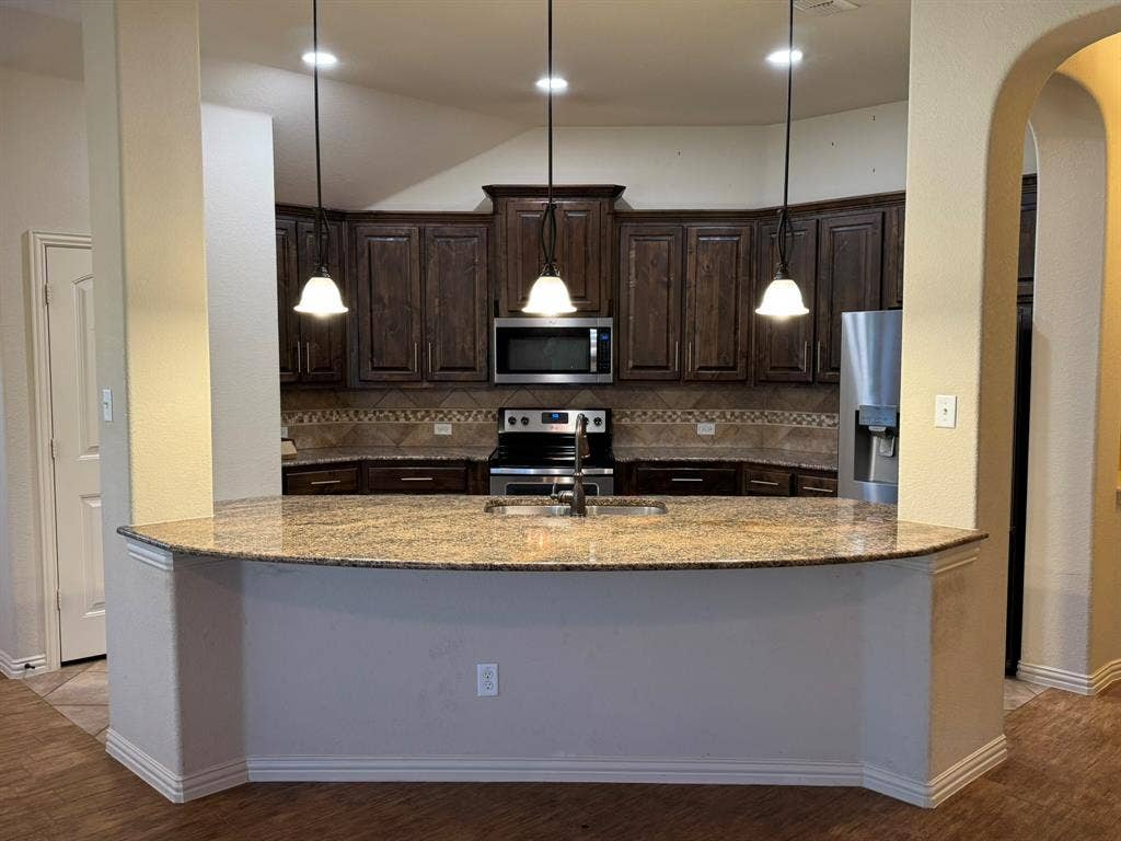 3953 Hollow Lake Road, Fort Worth, TX 76262 Kitchen featuring a curved stone-surface island, dark-toned wood cabinetry, built-in stainless steel appliances, decorative tile backsplash, and three pendant lights
