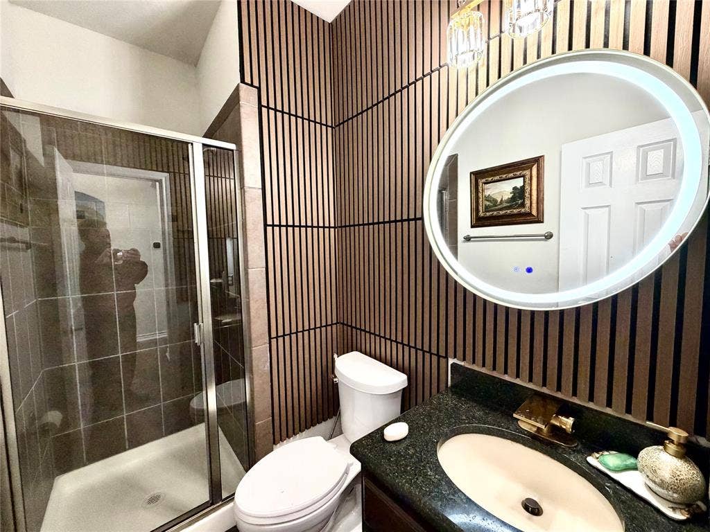 4514 Sea Sparrow Lane, Garland, TX 75043 Bathroom featuring vertical wood-slat wall paneling, an oval LED-backlit mirror, dark stone-finish countertop, integrated vanity sink with brass-tone faucet, and a glass-enclosed shower