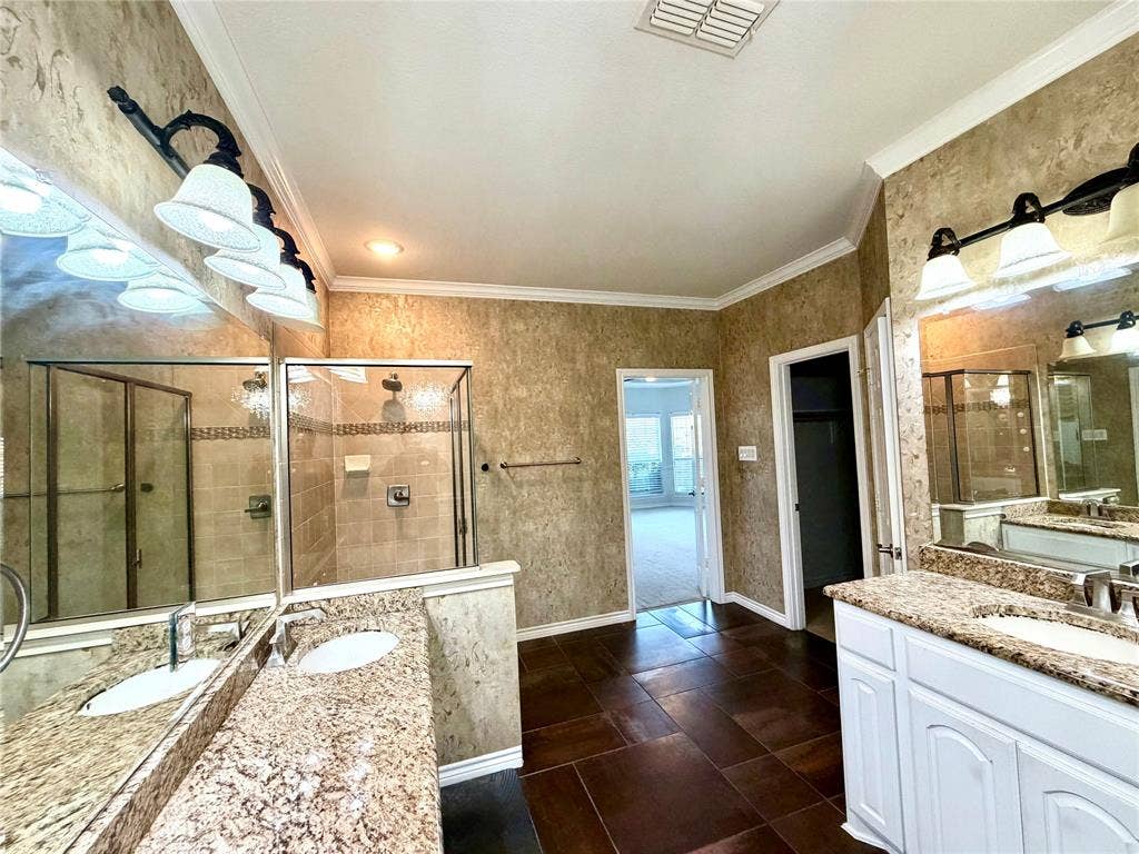 4514 Sea Sparrow Lane, Garland, TX 75043 Dual-vanity bathroom featuring stone-look countertops, white cabinetry, large plate glass mirrors with multi-light wall fixtures, and a glass-enclosed shower with tile surrounds, finished with oversized tile flooring