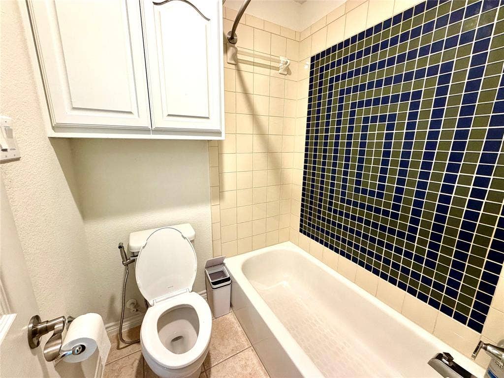 4514 Sea Sparrow Lane, Garland, TX 75043 Full bathroom featuring a tub-shower combination with geometric multi-colored wall tile, cream-toned square tile surround, built-in white upper cabinetry, and a high-profile commode with an attached handheld sprayer