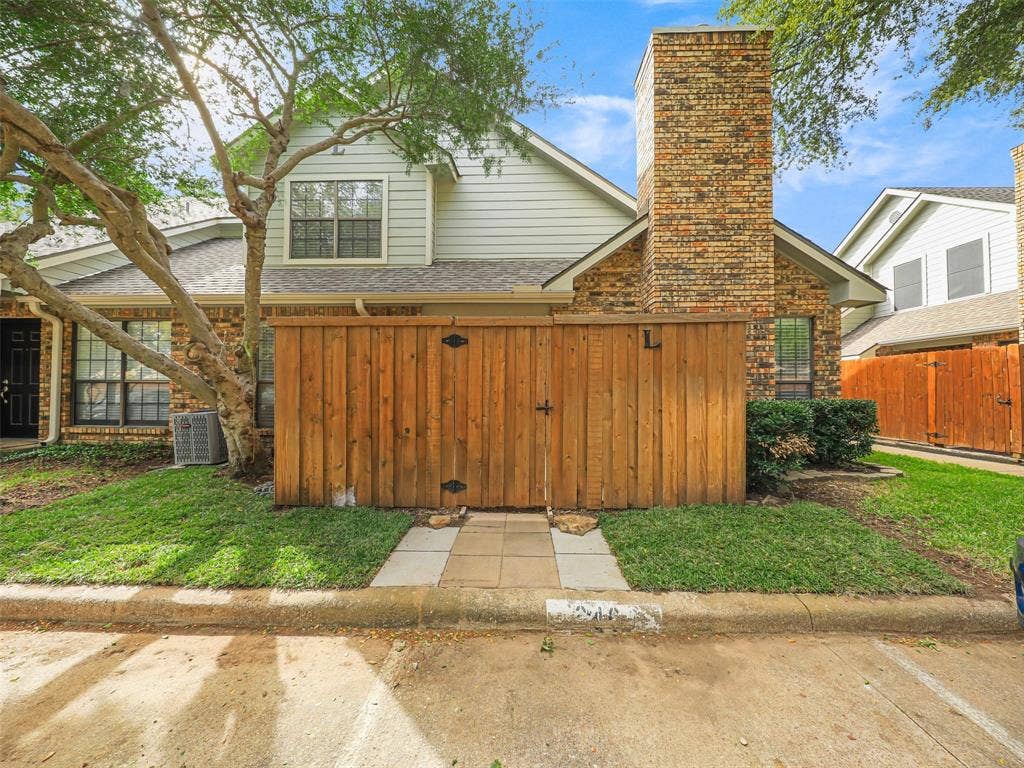 449 Harris Street, #102L, Coppell, TX 75019 Exterior elevation featuring a brick and horizontal siding facade, a prominent brick chimney, and a wooden privacy fence with a stone-paved walkway