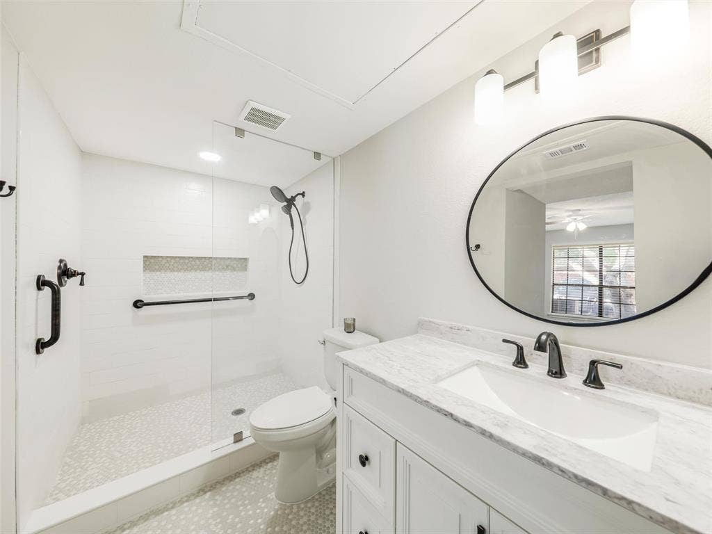 449 Harris Street, #102L, Coppell, TX 75019 White vanity with a stone countertop, oval mirror, walk-in shower with a glass panel, stone tile flooring, and matte black fixtures