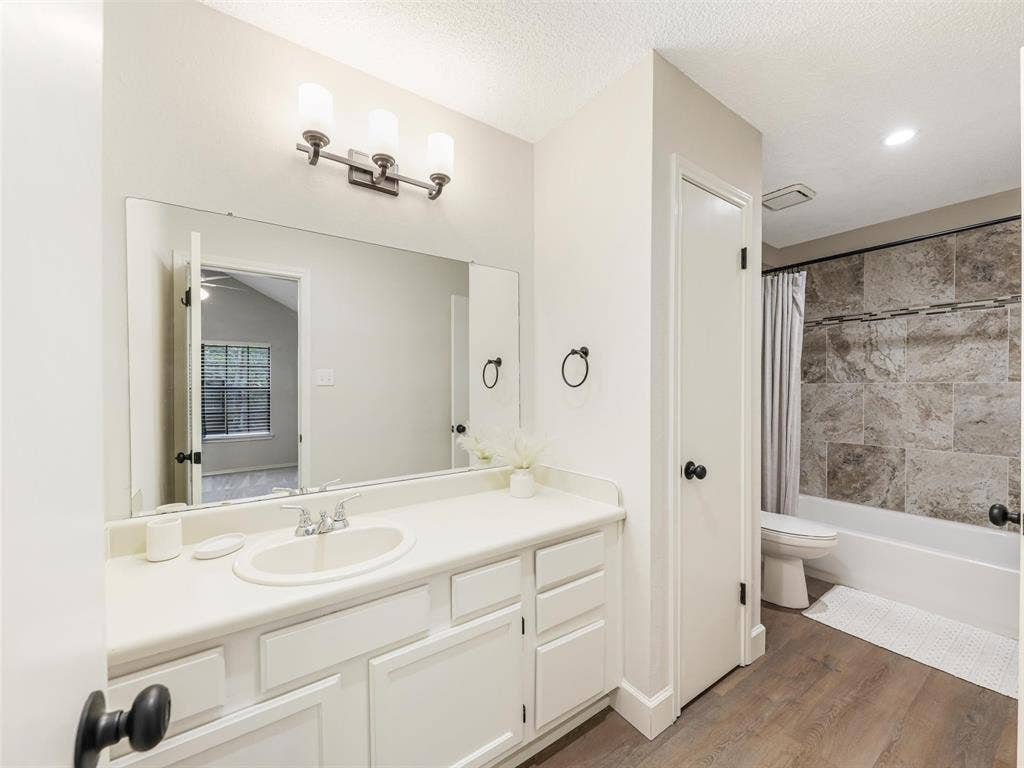 449 Harris Street, #102L, Coppell, TX 75019 White vanity featuring a molded sink and integrated countertop, full-width wall mirror with three-light fixture, wood-finish flooring, and bathtub surround accented with large-format stone-look tile