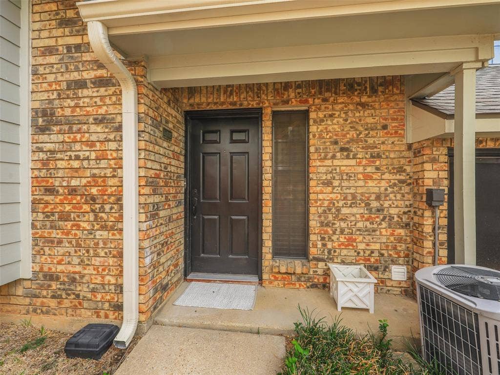 449 Harris Street, #102L, Coppell, TX 75019 Exterior entryway featuring a multi-tone brick facade, six-panel dark entry door, sidelight window, and a covered porch with concrete flooring