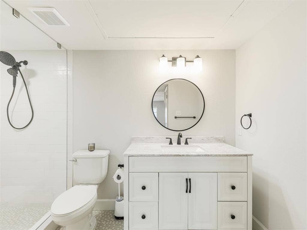 449 Harris Street, #102L, Coppell, TX 75019 Bathroom featuring a white vanity with stone-top surface, undermount sink, round wall-mounted mirror, three-light fixture, and a glass-enclosed shower with handheld sprayer