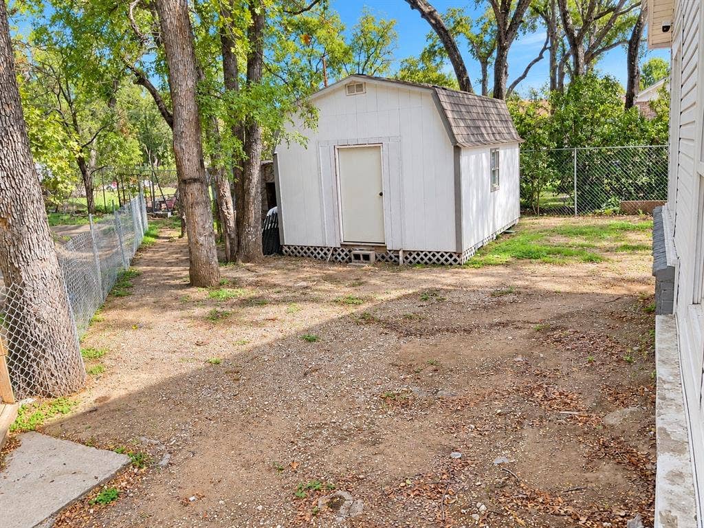 Detached storage shed with a gambrel roof, set within a fenced outdoor space featuring mature trees and lattice underpinning