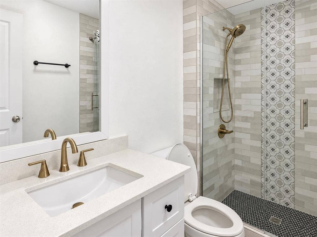 White vanity with light-toned countertop, integrated undermount sink, brass finish widespread faucet, glass-enclosed shower with decorative accent tiling, and brass finish handheld shower fixture