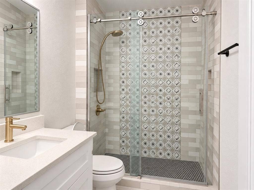 Bathroom featuring a white vanity with a neutral solid-surface countertop, gold-toned faucet, walk-in shower with sliding glass door, decorative patterned tile accent wall, and circular floor tile