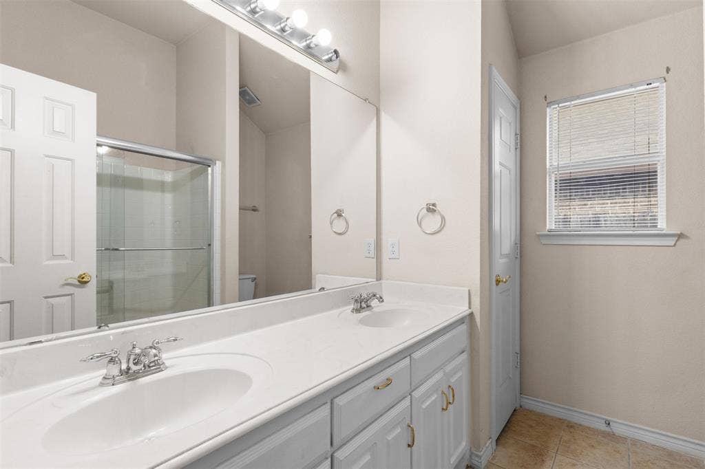 Double vanity featuring white cabinetry, integrated sinks with polished chrome fixtures, expansive wall-mounted mirror, and tile flooring