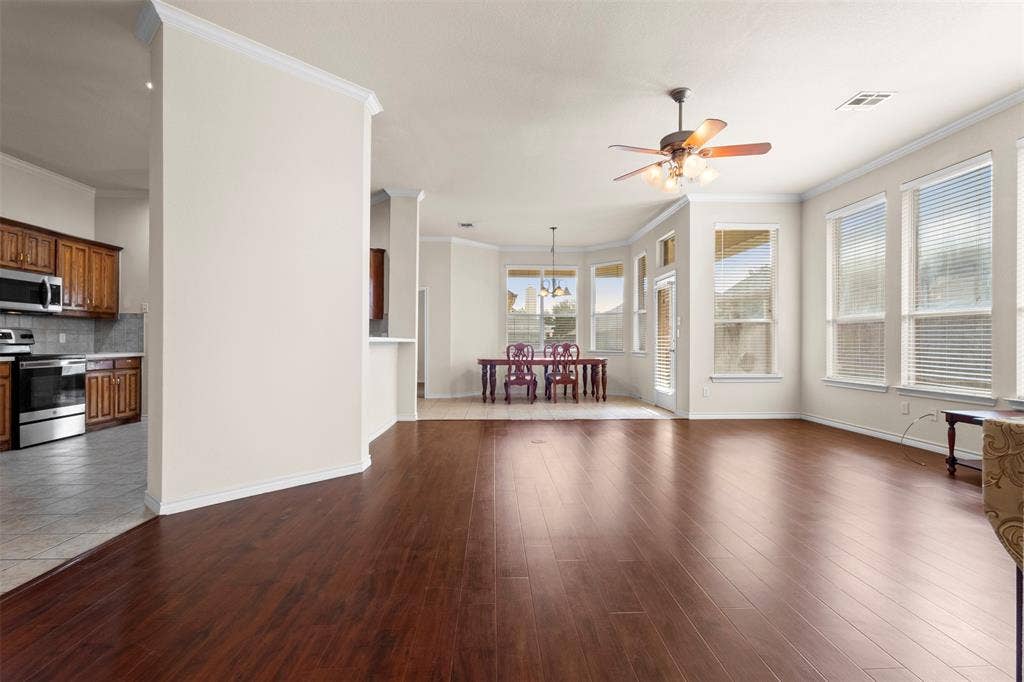 Open-concept interior featuring wood-finish flooring, multi-pane windows with blinds, crown molding, a ceiling fan with integrated lighting, and a kitchen with wood-finish cabinetry