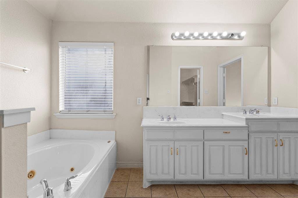 Dual-sink vanity with white cabinetry and stone-finish countertop, jetted soaking tub, oversized wall-mounted mirror, and tile flooring