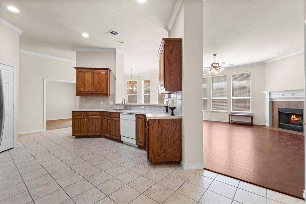Kitchen featuring wood cabinetry, neutral tile flooring, and integrated countertop space, complemented by a living area with wood-finish flooring and a tiled fireplace surround