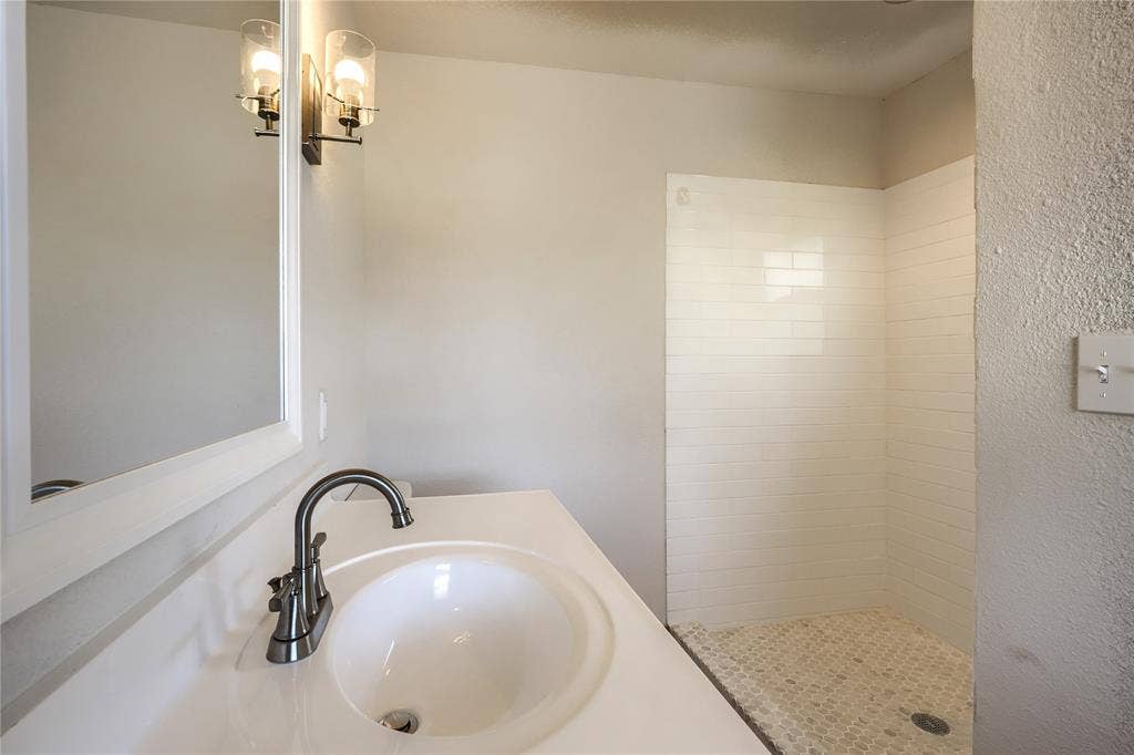 Bathroom vanity featuring a white countertop with an integrated oval sink, metallic gooseneck faucet, wall-mounted light fixture with glass shades, and a shower enclosure with white subway tile surround and mosaic floor tiling
