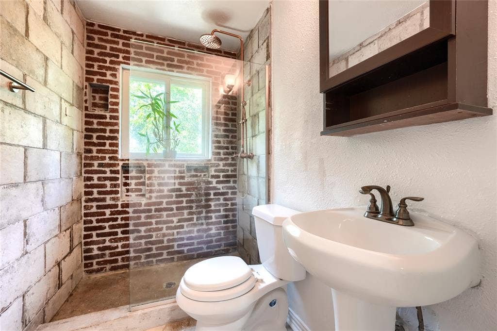 Walk-in shower featuring exposed brick walls, a glass partition, and a wall-mounted showerhead, accompanied by a pedestal sink with a metallic faucet and a built-in medicine cabinet