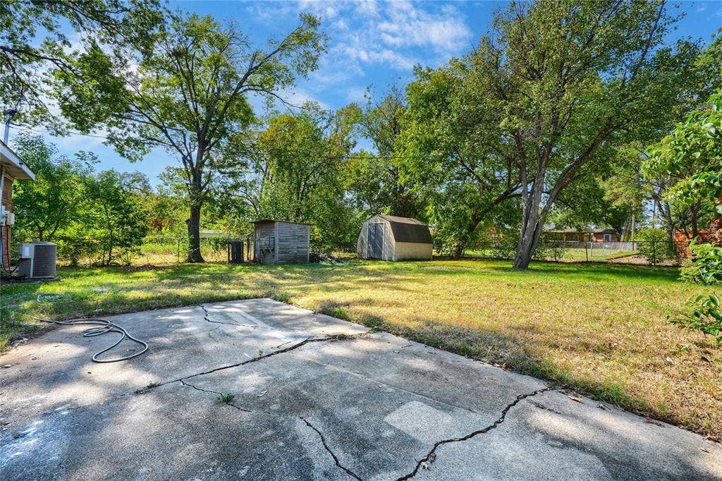 Backyard featuring a concrete patio, perimeter chain-link fencing, mature shade trees, and two storage structures