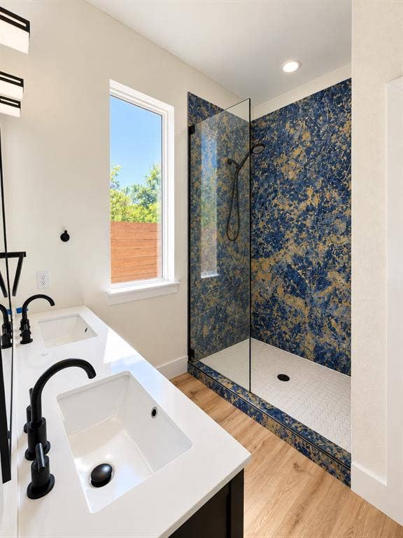 Dual-sink vanity with white countertops and matte black fixtures, walk-in shower featuring blue patterned tile surround and glass enclosure, and wood-finish flooring