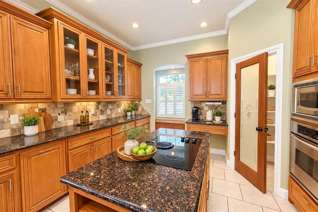 Kitchen featuring natural-finish cabinetry, stone countertops, glass-front upper cabinets, an integrated cooktop island, and a pantry with an etched-glass door