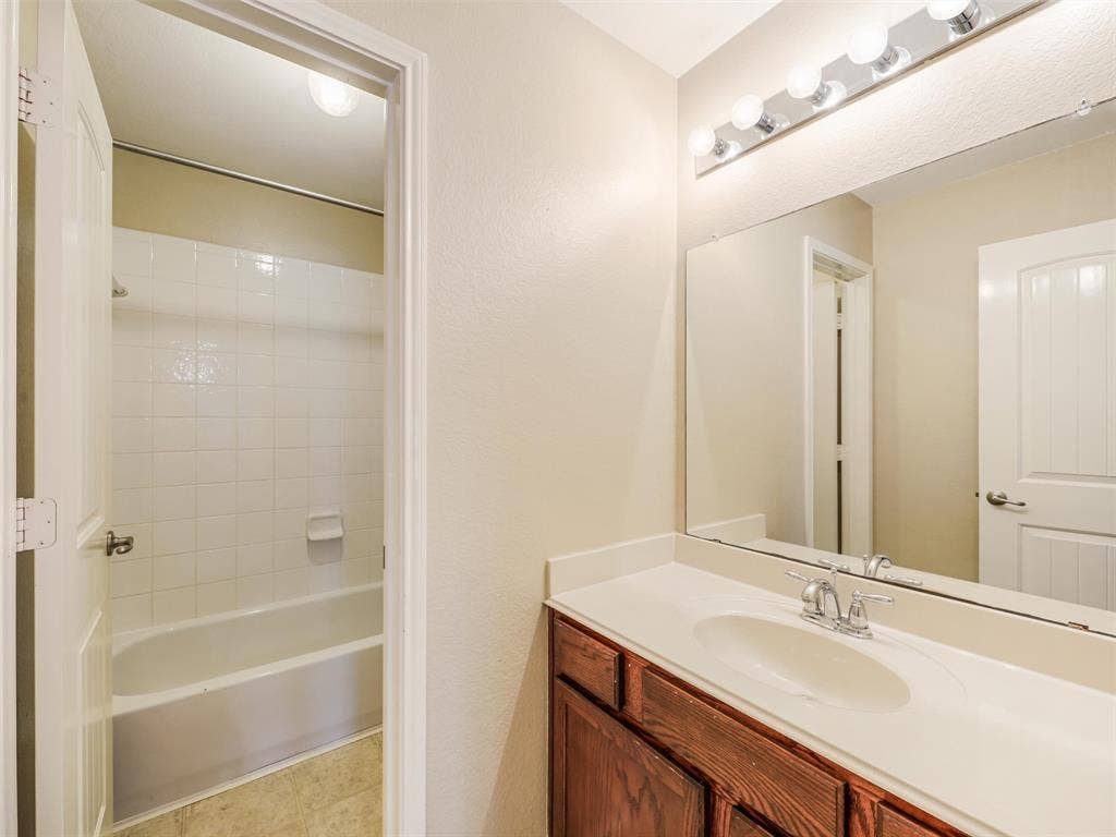 9720 Tipperary Drive, McKinney, TX 75072 Bathroom featuring a vanity with wood-finish cabinetry, a white integrated countertop and basin, a full-width mirror, and a wall-mounted light fixture, with a separate tub and shower enclosure tiled in white ceramic