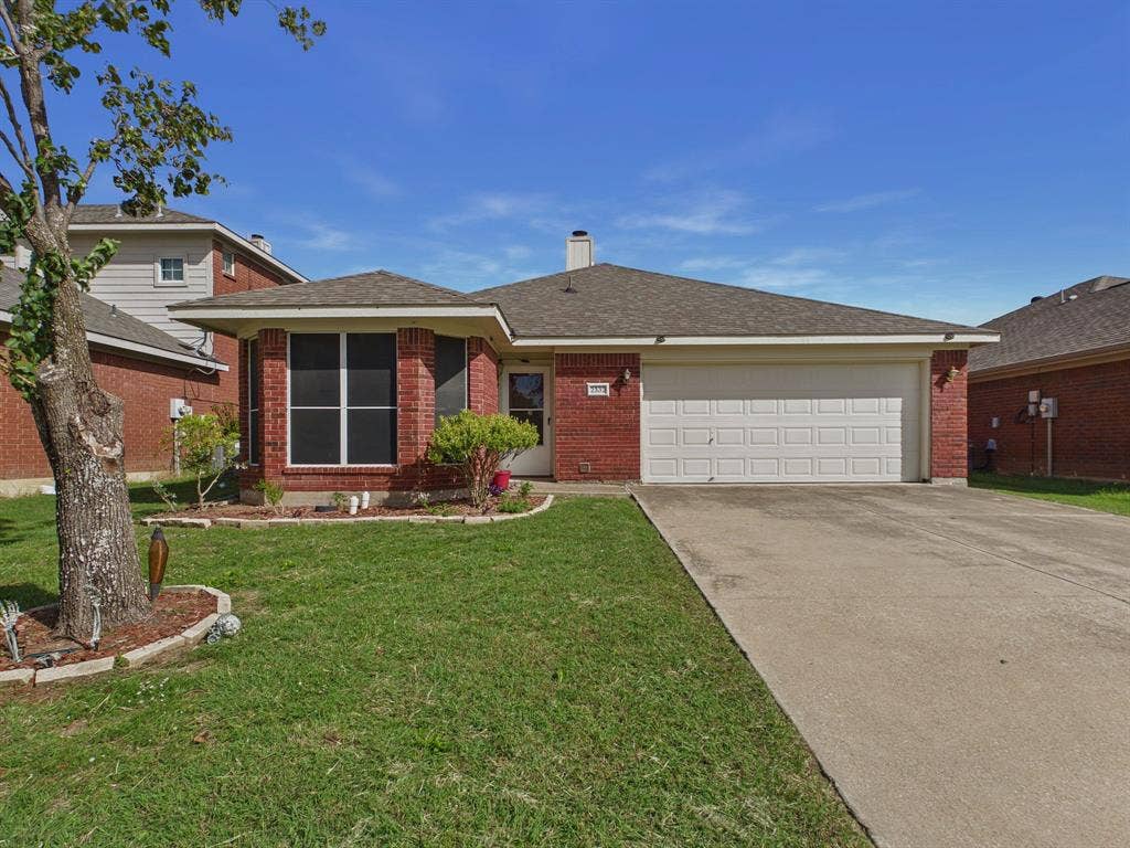 Single-story residence featuring a brick exterior, shingled roof, attached two-car garage, concrete driveway, and a front garden bed with stone edging