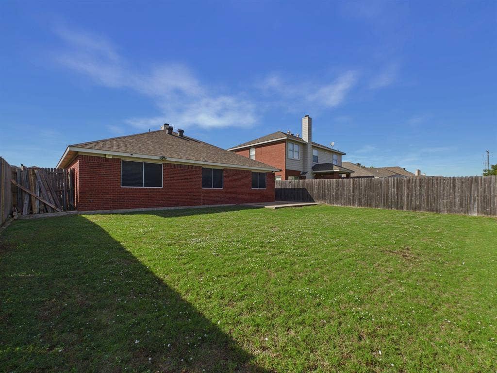 Rear exterior featuring a brick facade, hip roof with composition shingles, concrete patio, and fenced-in backyard with a grass lawn