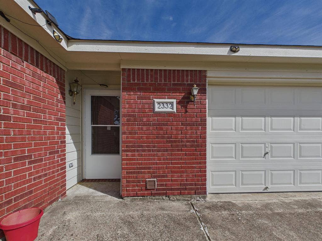 Red brick facade with a white, paneled garage door, recessed entryway, and exterior wall-mounted lighting