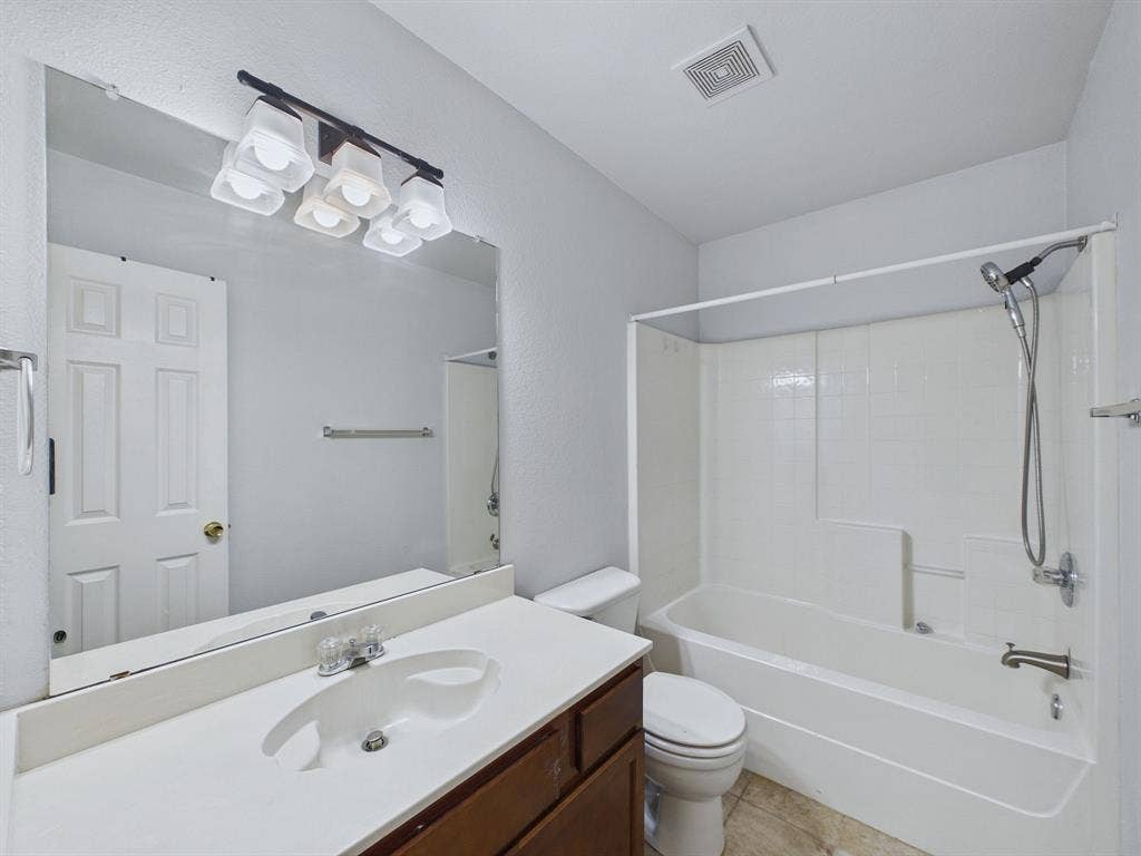 Full-width vanity with white solid-surface countertop and integrated sink, chrome multi-light fixture, oversized frameless mirror, and shower-tub combination with a handheld showerhead