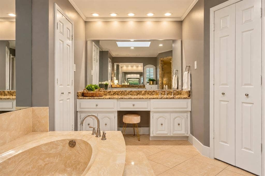 5833 Gallant Fox Lane, Plano, TX 75093 Custom vanity featuring white cabinetry, stone countertop, and integrated makeup station, complemented by a stone-surround soaking tub, large wall-mounted mirror, recessed lighting, and tile flooring