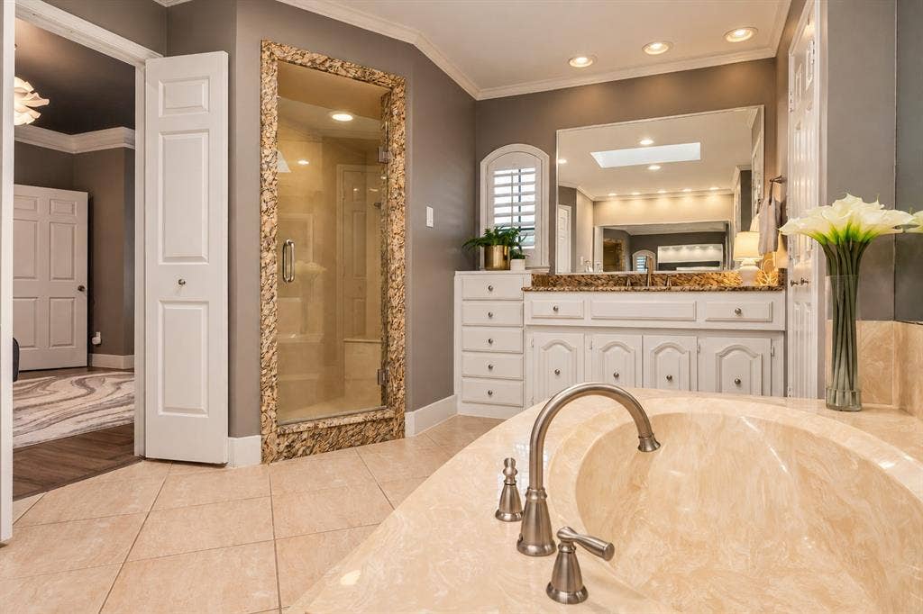 5833 Gallant Fox Lane, Plano, TX 75093 Bathroom featuring an oval soaking tub with a stone-finish deck, a custom-tiled walk-in shower with a stone surround, white cabinetry, a large wall-mounted mirror, and tile flooring