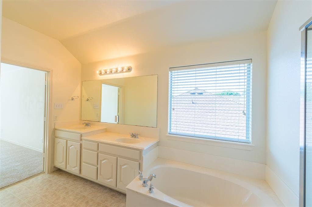 Bathroom featuring a dual-sink vanity with white cabinetry, a large wall-mounted vanity mirror, a soaking tub with deck-mounted chrome fixtures, and a window with horizontal blinds