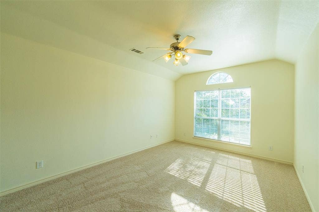 Vaulted ceiling featuring a central multi-blade ceiling fan with integrated lighting, neutral carpeted flooring, and a large window topped with an arched transom