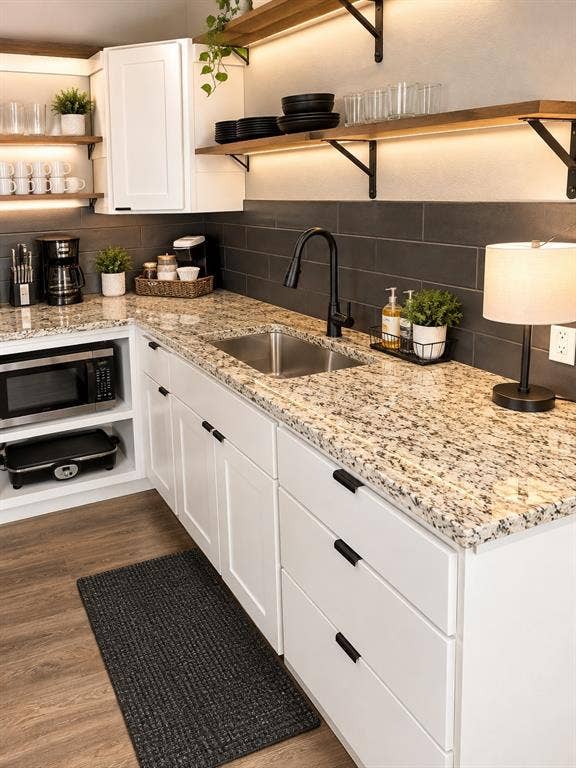 1579 Eastside Lake Road, Graham, TX 76450 Kitchen featuring white cabinetry with matte black hardware, stone-finish countertops, dark rectangular tile backsplash, wood-finish shelving with under-cabinet lighting, and a stainless steel undermount sink with a matte black faucet