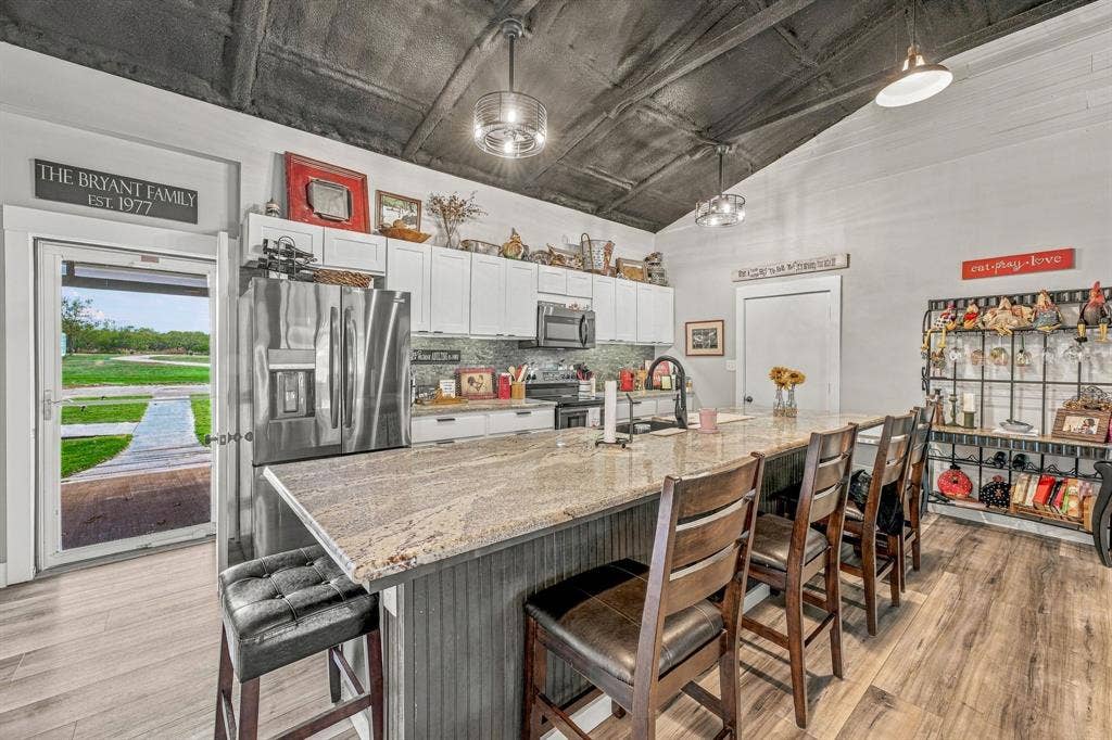 1579 Eastside Lake Road, Graham, TX 76450 Kitchen featuring wood-finish flooring, granite-topped center island with vertical slat cabinetry, white upper and lower cabinetry, stainless steel appliances, and vaulted ceiling with exposed rafters