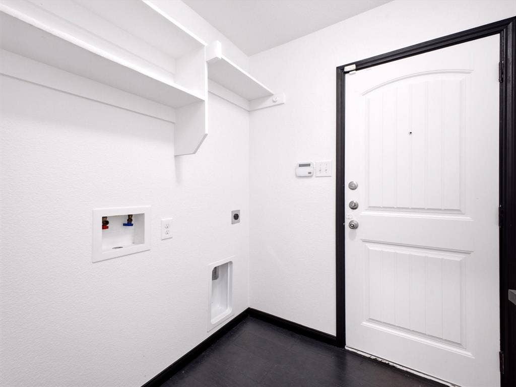 Utility area featuring white built-in wall shelving, dedicated water hookups, dark flooring, and a solid-core door with a high-contrast trim