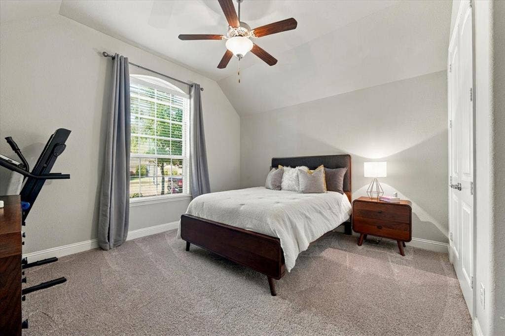 1455 Corrara Drive, Rockwall, TX 75032 Suite Bedroom features a vaulted ceiling and a ceiling fan with light fixture, and an arched-top window overlooking the front yard. Carpet flooring extends throughout.