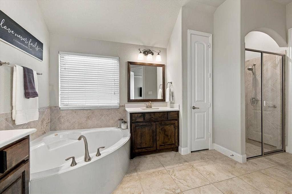 1455 Corrara Drive, Rockwall, TX 75032 Primary Bathroom features a vaulted ceiling, a vanity light fixture, a corner garden tub with tile surround beneath a window, a linen closet, and a separate walk-in shower with glass enclosure set within an arched opening. Ceramic tile flooring extends throughout.