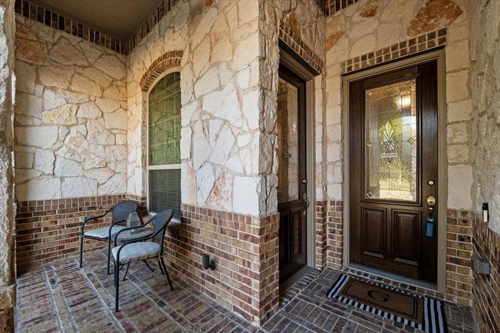 1455 Corrara Drive, Rockwall, TX 75032 Covered front porch features stone and brick walls, an arched window, a front entry door with decorative glass, and a separate exterior entry to the suite sitting room. Brick flooring extends throughout.
