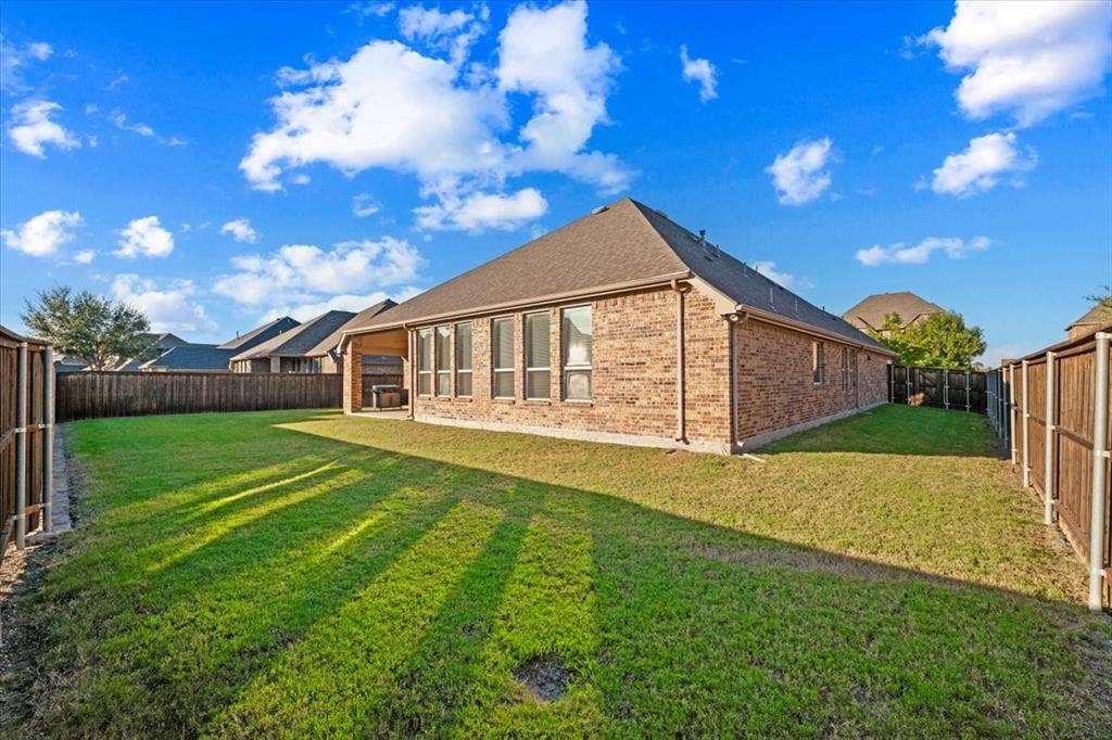1455 Corrara Drive, Rockwall, TX 75032 Backyard features a view of the hip roofline and brick construction with gutters, a row of windows along the back of the home, wood privacy fencing with metal posts, and an open grass lawn.