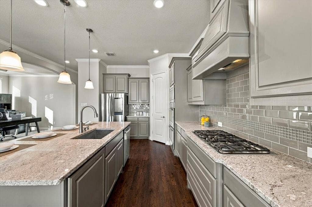 1455 Corrara Drive, Rockwall, TX 75032 Kitchen features recessed lighting, a subway tile backsplash with a mosaic accent band, a gas cooktop, an island with an undermount sink and stainless dishwasher, a pantry, and an arched opening to the office. Wood flooring extends throughout.