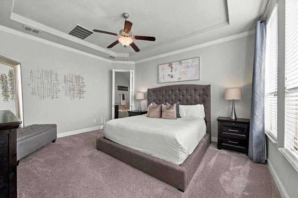 1455 Corrara Drive, Rockwall, TX 75032 Primary Bedroom features a tray ceiling with crown molding and a ceiling fan with light fixture, a wall of side windows, and a doorway connecting to the primary bathroom. Carpet flooring extends throughout.