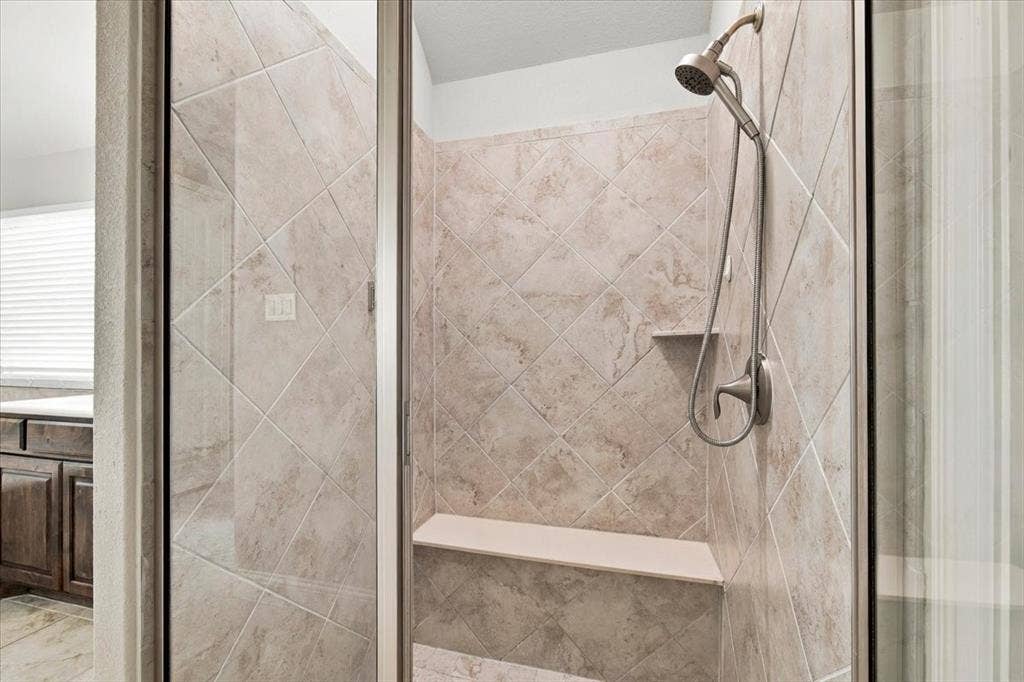 1455 Corrara Drive, Rockwall, TX 75032 Primary Bathroom features a walk-in shower with a glass enclosure, diagonal tile surround, a built-in bench, a corner shelf, and a handheld showerhead. Ceramic tile flooring extends throughout.