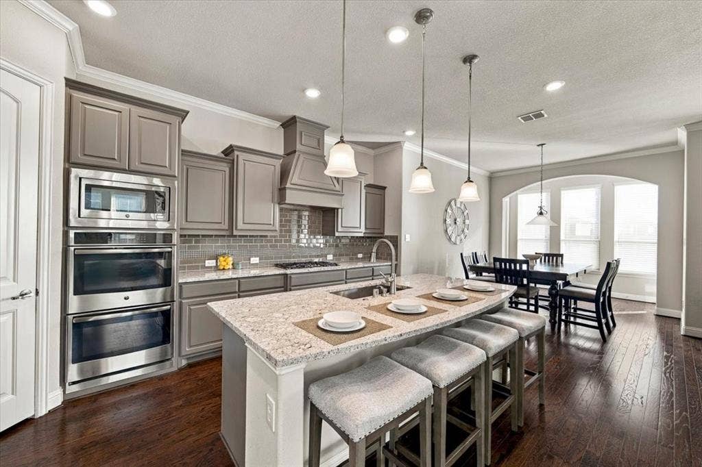 1455 Corrara Drive, Rockwall, TX 75032 Kitchen features gray cabinetry, a built-in stainless microwave and double wall oven, countertops along the perimeter and island, and an open layout to the dining area. Wood flooring extends throughout.