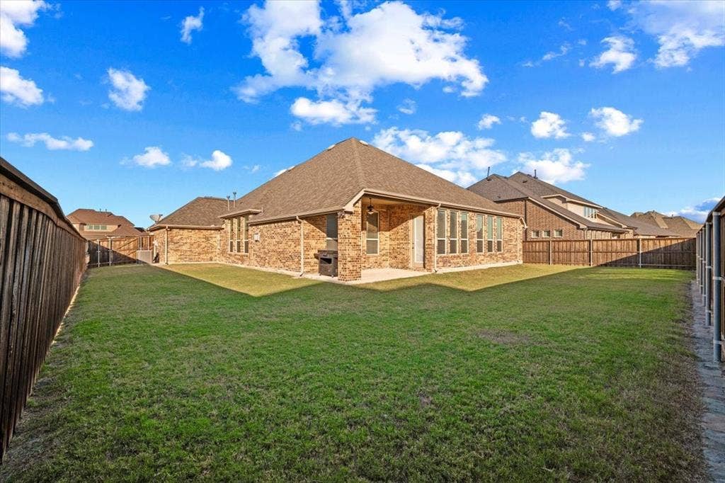 1455 Corrara Drive, Rockwall, TX 75032 Backyard features a view of the composition roof and brick construction, a covered patio, multiple windows, wood privacy fencing with metal posts, an underground sprinkler system, and a spacious grass lawn.