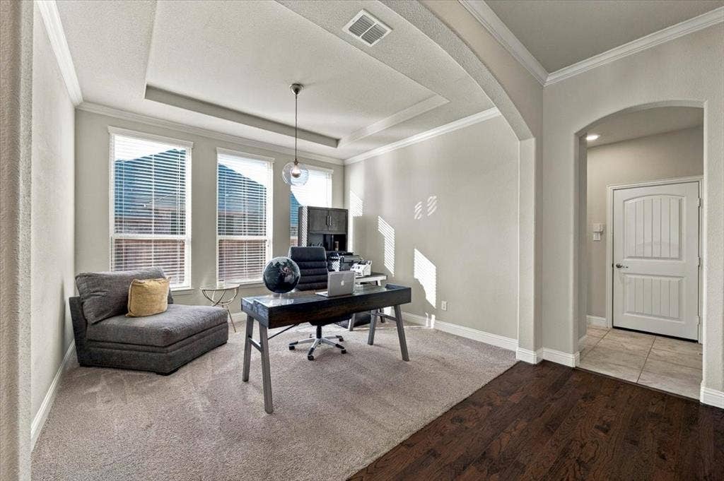 1455 Corrara Drive, Rockwall, TX 75032 Office features a tray ceiling with crown molding, a pendant light fixture, three windows, and arched openings to the main hall and the adjacent hall. Carpet flooring extends throughout, transitioning to wood and ceramic tile in the adjoining halls.