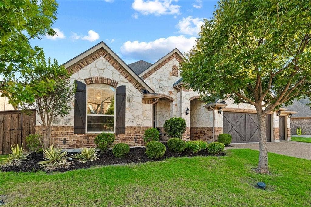1455 Corrara Drive, Rockwall, TX 75032 Front exterior features gabled rooflines with brick accents, stone and brick construction, an arched window with shutters, stone archways leading to the covered entry, wood privacy fencing, mature trees, landscaped beds, and an underground sprinkler system.