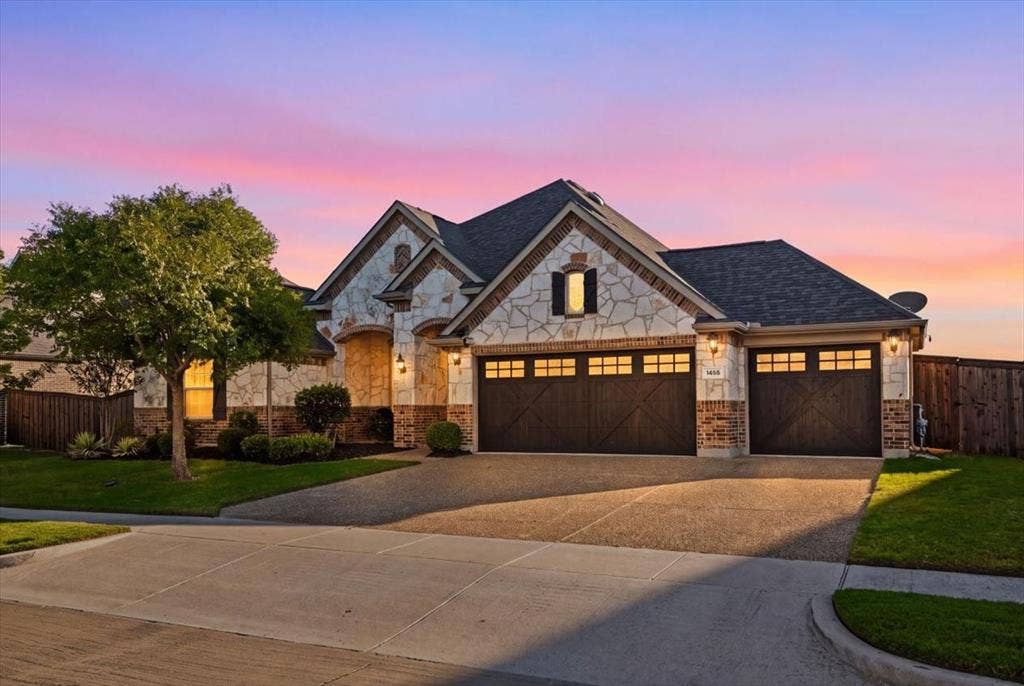 1455 Corrara Drive, Rockwall, TX 75032 Front exterior features a composition roof with gabled rooflines, stone and brick construction, coach lights, an arched window with shutters, an attached three-car garage with carriage-style doors, a mature tree, landscaped beds, and a concrete driveway.