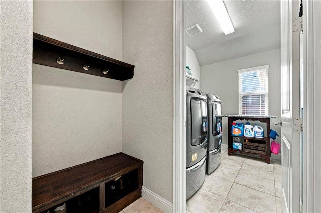 1455 Corrara Drive, Rockwall, TX 75032 Laundry Room features an angled ceiling section, an overhead light fixture, a wall-mounted shelf, a window, and a built-in drop zone outside the entry with a wall-mounted hook rail and a bench with storage cubbies. Ceramic tile flooring extends throughout.