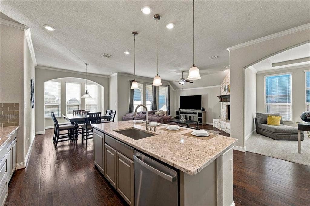 1455 Corrara Drive, Rockwall, TX 75032 Kitchen features a center island with an undermount sink overlooking the living room, an arched opening to the office, and an open layout to the dining area. Wood flooring extends throughout, transitioning to carpet in the office.