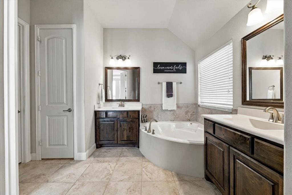 1455 Corrara Drive, Rockwall, TX 75032 Primary Bathroom features a vaulted ceiling, vanity light fixtures, separate vanities with dark wood cabinetry, a corner garden tub with tile surround beneath a window, and a linen closet. Ceramic tile flooring extends throughout.