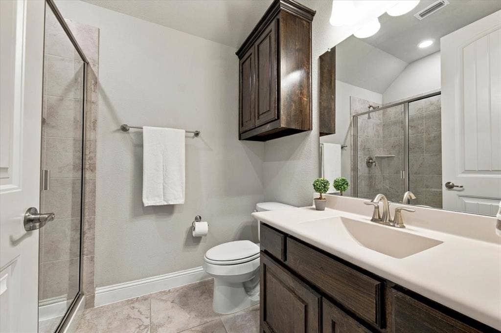 1455 Corrara Drive, Rockwall, TX 75032 Suite Bathroom features a vanity light fixture, a cabinet above the toilet, a single vanity with countertop and cabinet storage, and a walk-in shower with a glass door and tile surround. Ceramic tile flooring extends throughout.